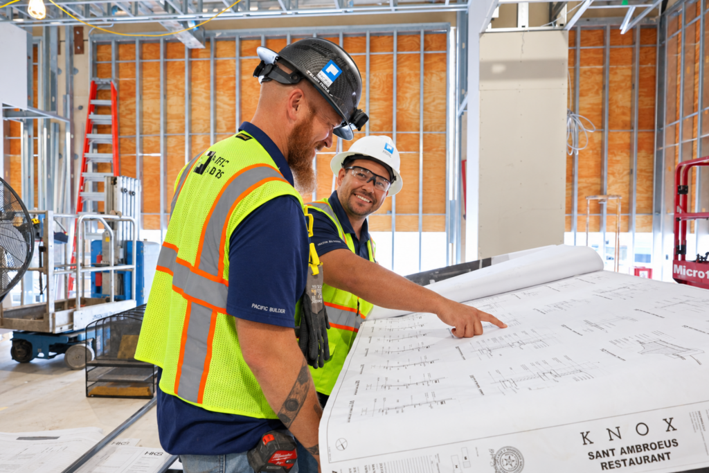 Two Pacific Builders team members in hard hats and safety vests reviewing construction plans on a jobsite